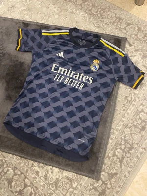 Real Madrid bortatröja Vini Jr. 7 - Mörkblå Real Madrid bortatröja från Adidas med Vini Jr. och nummer 7 på ryggen. Tröjan har mönstrad design, gula detaljer på ärmar och axlar samt klubbmärke och sponsortryck framtill. Tillverkad i lätt och ventilerande material.