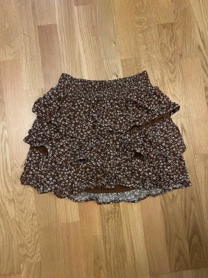 Brun blommig volangkjol från Lindex - Söt brun kjol från Lindex med små vita blommor och flera volanglager. Kjolen har resår i midjan för en bekväm passform. Barnstorlek 146/152, sällan använd. 