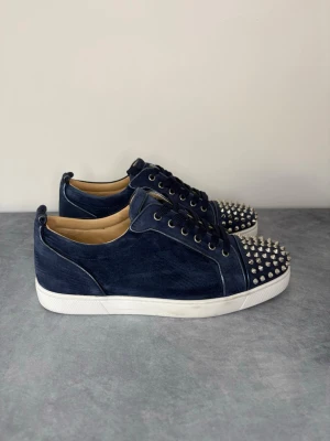 Blåa Louboutin sneakers med nitar - Mörkblå sneakers i mocka från Christian Louboutin med coola silverfärgade nitar på tån. Skorna har vita sulor, svarta skosnören och en klassisk låg profil. Insidan är beige och märkt med Louboutin-loggan. Perfekta för dig som vill sticka ut med din stil.
