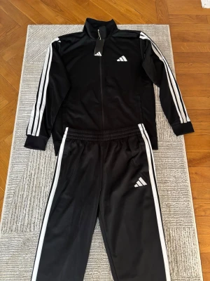 Svart Adidas träningsset med vita ränder - Snyggt svart träningsset från Adidas med klassiska vita ränder längs ärmar och ben. Setet består av en zip-jacka med hög krage och matchande byxor med resår i midjan. Båda plaggen har Adidas-loggan tryckt på bröstet och benet. Perfekt för träning eller chill. ( PRIS KAN DISKUTERAS )