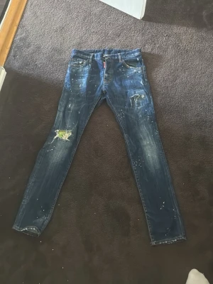 Dsquared2 blå jeans med färgstänk - Säljer ett par blå jeans från Dsquared2 med coola färgstänk och slitningar. Jeansen har raka ben och klassisk femficksdesign. Detaljer som patch och spruckna partier ger ett edgy uttryck. Perfekt för dig som gillar streetstyle och unika plagg. Finns en reva som absolut går o laga hur vilken skräddare som helst om man vill