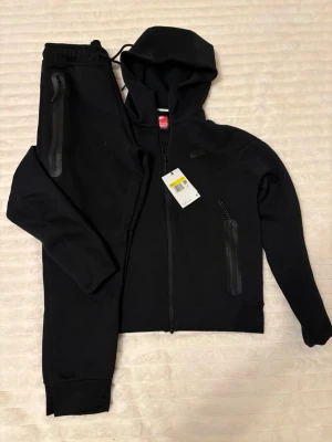 Svart Nike loungewear set med huva - Snyggt svart loungewear set från Nike med zip hoodie och matchande byxor. Setet har dragkedjor på fickorna och en sportig look. Perfekt för chill dagar hemma eller när du vill vara bekväm men ändå se fräsch ut.