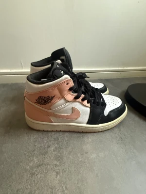 Air Jordan 1 Mid rosa/svart/vit - Snygga Air Jordan 1 Mid sneakers i rosa, svart och vitt. Skorna har klassisk Jordan-logga på sidan, svart snörning och vit sula. Ovandelen är i läder med perforerad tåbox för extra ventilation. Perfekt för dig som gillar streetwear och vill sticka ut.