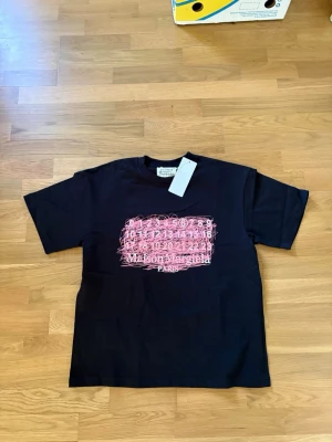 Svart Maison Margiela t-shirt - Cool svart t-shirt från Maison Margiela med ett unikt rosa grafiskt tryck framtill. Klassisk passform med korta ärmar och rund halsringning. Perfekt statement-plagg för dig som gillar designerkläder och streetstyle.
