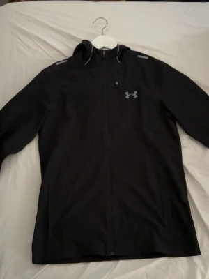 Svart vindjacka från Under Armour - Snygg svart vindjacka från Under Armour med huva och dragkedja framtill. Jackan har en diskret logga på bröstet och reflekterande detaljer på axlarna. Perfekt för blåsiga dagar och sportiga outfits.