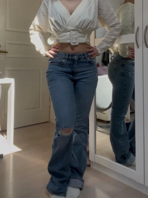 bootcut jeans  - Snygga blå bootcut jeans med klassisk femficksdesign och ett slitande hål på ena knät. Jeansen har hög midja och är tillverkade i denim med lätt stretch för skön passform. Perfekta för en trendig och avslappnad look.