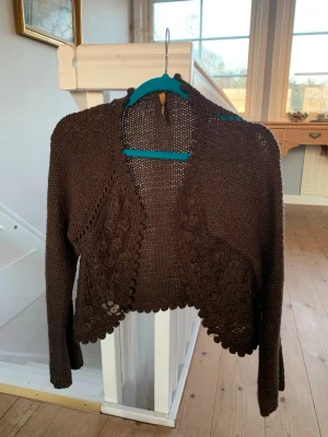 Brun stickad bolero med spetskant - Mysig brun stickad bolero med lång ärm och dekorativ spetskant. Kortare modell med öppet framtill och fina virkade detaljer längs kanterna. Perfekt att slänga över en topp för en extra touch. Materialet känns mjukt och följsamt.