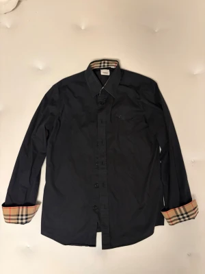 Svart skjorta från Burberry, M - Stilren svart skjorta från Burberry med klassiskt rutigt mönster på insidan av kragen och ärmsluten. Skjortan har långa ärmar, knappar framtill och är tillverkad i bomull. Perfekt för dig som gillar exklusiva detaljer och tidlös design.