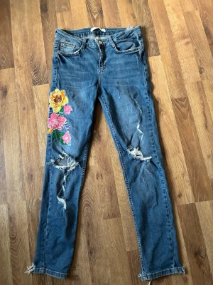 Blå jeans med blommor och slitningar - Säljer ett par blå skinny jeans med färgglada broderade blommor och slitningar på båda benen. Jeansen har klassisk femficksmodell