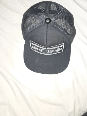 Svart Zara keps - Snygg svart keps från Chrome Hearts med vit broderad patch framtill där det står 'Chrome Hearts Hollywood, USA' och CH-logga. Kepsen har mesh-bakstycke för extra ventilation och justerbar passform. Perfekt för en streetwear-look.