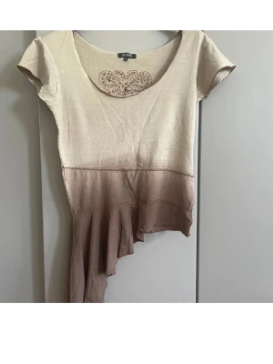 Beige och brun asymmetrisk topp Sisters - Unik topp från Sisters i beige och brun med asymmetrisk volangkant. Kortärmad modell med rund halsringning och spetsbroderat hjärta fram och bak. Perfekt för dig som vill sticka ut med en kreativ och cool stil.