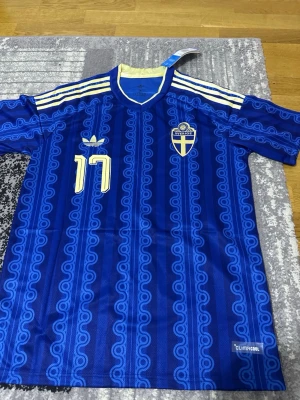 Blå Sverige matchtröja Adidas - Säljer en blå Sverige matchtröja från Adidas med nummer 17 på bröstet. Tröjan har gula detaljer, vita ränder på axlarna och ett retroinspirerat mönster. Tillverkad i lätt och ventilerande material med ClimaCool-teknologi. Perfekt för fotboll eller samling.