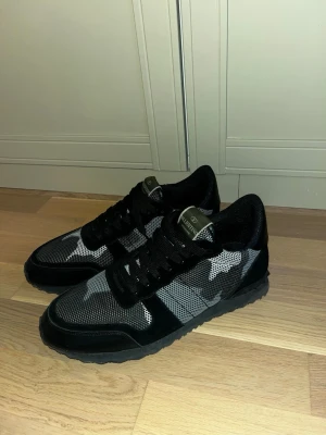 Svarta Valentino sneakers med kamomönster - Snygga svarta sneakers från Valentino med grått och vitt kamouflagemönster. Skorna har svarta snören, mesh- och mockadetaljer samt en robust svart sula. Perfekta för dig som vill sticka ut med en exklusiv och trendig look.