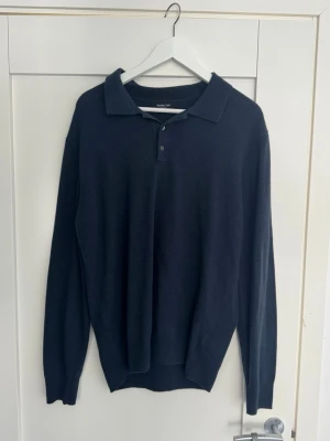 Mörkblå pikétröja från Massimo Dutti - Stilren mörkblå långärmad pikétröja från Massimo Dutti med klassisk krage och två knappar framtill. Tröjan har en smal passform och är tillverkad i ett mjukt, tunt material som känns skönt mot huden.
