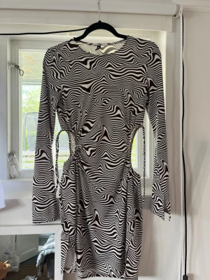Zebramönstrad kort klänning med cut-outs - Snygg kort klänning med svartvitt zebramönster och långa ärmar. Klänningen har trendiga cut-outs i midjan och figurnära passform. Perfekt för dig som vill sticka ut med ett coolt mönster och unika detaljer. Anpassningsbar storlek då man kan göra cut outsen större eller mindre. 