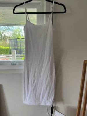 Vit underklänning i lite tjockare material  - Säljer en enkel och stilren vit klänning med smala axelband och rak passform. Klänningen är ärmlös och slutar ovanför knäna, perfekt för varma dagar. Materialet känns mjukt och lätt mot huden.