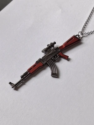 Halsband med gevärshänge i metall - Unikt halsband med ett detaljerat gevärshänge AK47 i metall. Hänget har inslag av brun och silverfärg, med realistiska detaljer som magasin och sikte. Perfekt accessoar för dig som gillar edgy och annorlunda smycken. Materialet rostfritt stål. 