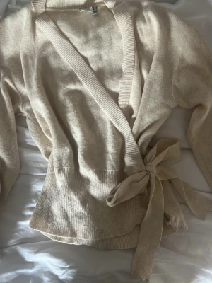 omlottkofta - Snygg beige omlottkofta med breda ribbade kanter och knytband i midjan. Koftan har lång ärm och är tillverkad i ett mjukt, stickat material som ger en avslappnad men stilren look.