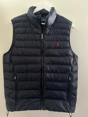 Svart dunväst från Polo Ralph Lauren - Snygg svart dunväst från Polo Ralph Lauren med quiltad design. Västen har dragkedja framtill, två fickor med dragkedja och den klassiska röda Polo-loggan broderad på bröstet. Perfekt för sommarn/ hösten nästan aldrig använd 