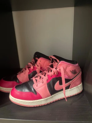 Nike Air Jordan 1 Mid rosa/svart - Säljer ett par Nike Air Jordan 1 Mid sneakers i rosa och svart läder med vita sulor. Klassisk siluett med ikonisk Swoosh och Air Jordan-logga på sidan. Skorna har rosa snören och kontrasterande paneler för en riktigt snygg look.