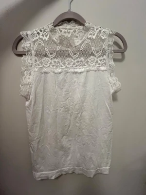 Vit spetsdetaljerad ärmlös topp - Söt vit topp med spets upptill och runt axlarna. Ärmlös modell med rund halsringning och fina blommiga spetsdetaljer. Perfekt att styla med jeans eller kjol för en fräsch look. Köpt från någon liten butik i Skåne men knappt använd 💕