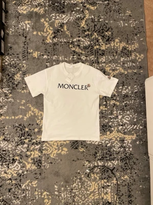 Vit Moncler t-shirt med logga - Snygg vit t-shirt från Moncler med klassisk svart logotyp på bröstet. T-shirten har rund halsringning och korta ärmar. Perfekt för en stilren och trendig look.