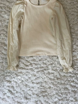 Beige blus med puffärm och struktur - Stilren beige blus med puffiga långärmar och diskreta ränder i tyget. Blusen har rund halsringning och små volangdetaljer vid ärmsluten. Använd en gång och är dom ny. Strl 146/152