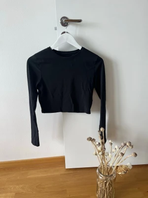 Svart croppad långärmad topp 157 - Svart croppad långärmad topp från 157. Enkel och stilren design med rund halsringning. Tillverkad i mjuk bomull med lite stretch för en bekväm passform. Perfekt att matcha med höga jeans eller kjol.