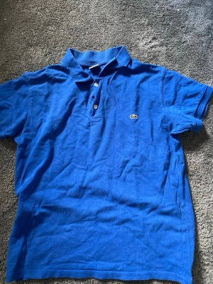 Blå pikétröja från Lacoste - Klassisk blå pikétröja från Lacoste med korta ärmar och två knappar framtill. Tröjan har den ikoniska krokodilloggan broderad på bröstet och är tillverkad i mjuk bomull. Perfekt till en avslappnad och stilren look.