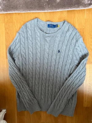 Grå kabelstickad tröja Polo Ralph Lauren - Kabelstickad grå tröja från Polo Ralph Lauren med rund halsringning och broderad marinblå logga på bröstet. Tröjan har långa ärmar och ribbade muddar vid ärmslut och nederkant. Perfekt för lager-på-lager och klassisk stil.