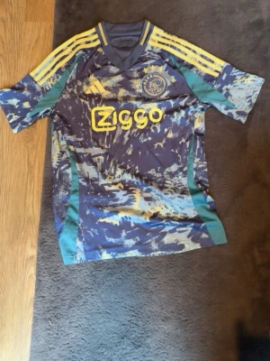Ajax matchtröja Adidas blå/gul - Ajax matchtröja från Adidas med blått och gult abstrakt mönster, gula ränder på axlarna och Ziggo-logga på bröstet. Klubblogga på vänster bröst och tre gula kryss i nacken. Tillverkad i lätt funktionsmaterial Aeroready.