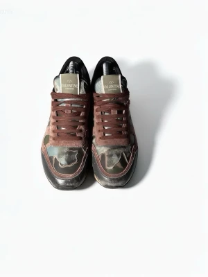Valentino camo sneakers med bruna detaljer - Snygga sneakers från Valentino med camo-mönster i grönt och brunt, bruna skosnören och svarta detaljer. Märkeslogga på plösen och klassisk låg siluett. Perfekta för dig som vill sticka ut med en exklusiv streetstyle-look.