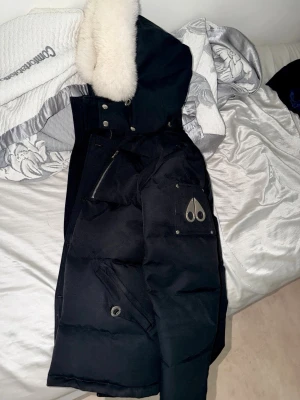 Svart dunjacka Moose Knuckles - Snygg svart dunjacka från Moose Knuckles med pälskantad huva och klassisk logga på ärmen. Jackan har flera fickor med dragkedja och tryckknappar, samt en rejäl och varm design perfekt för kalla vinterdagar.
