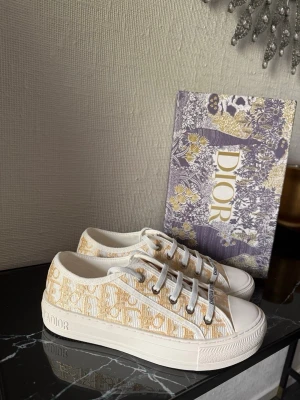 Dior Oblique sneakers i beige och vitt - Snygga Dior Oblique sneakers i beige och vitt med klassiskt monogrammönster. Skorna har vita snören, gummisula med Dior-logga och detaljer i canvas. Perfekta för dig som vill ha en lyxig och trendig look med tydlig designerstil.