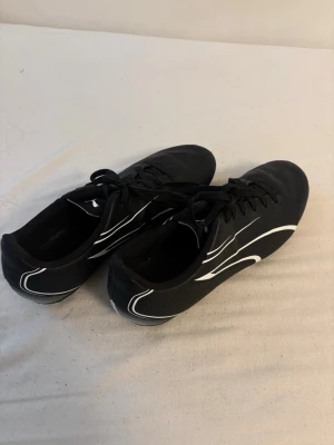 Svarta fotbollsskor från Puma - Snygga svarta fotbollsskor från Puma med vita detaljer och klassisk design. Skorna har snörning och är tillverkade i syntetmaterial som ger bra känsla på planen. Perfekta för dig som vill ha stilrena och funktionella skor till match eller träning.