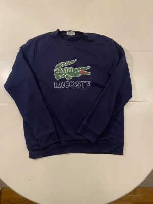 Mörkblå sweatshirt från Lacoste storlek M  - Mörkblå Lacoste tröja med den ikoniska krokodillogotypen broderad på framsidan.   Storlek: M  Längd: 65 cm  Armhålsbredd: 60 cm   Ställ gärna frågor! :)