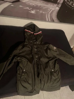Grön Moncler vindjacka med huva - Snygg grön vindjacka från Moncler med huva och dragkedja. Jackan har två fickor med knappar och en rödvit rand på huvan. Perfekt för blåsiga dagar och enkel att matcha med streetwear.