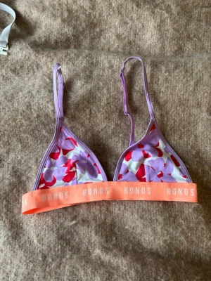 Lila och röd blommig bralette Bonds - Triangel-bh från Bonds i lila och rött med blommigt mönster. Justerbara axelband och bred orange resår med logga. Mjuk och bekväm modell utan bygel, perfekt för en avslappnad stil. ingen push up alls