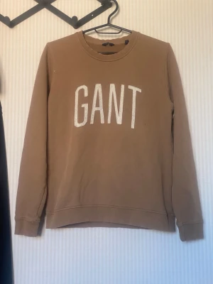 Beige sweatshirt från GANT - Säljer en beige sweatshirt från GANT med klassisk rund hals och stora vita bokstäver på bröstet. Tröjan har långa ärmar och ribbade muddar vid ärmslut och nederkant. Perfekt för dig som gillar stilrena och bekväma plagg.