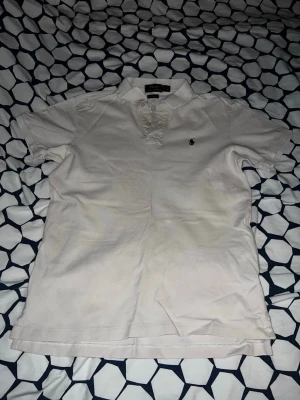 Vit pikétröja från Ralph Lauren - Klassisk vit pikétröja från Ralph Lauren med korta ärmar och liten broderad logga på bröstet. Tröjan har krage och knappar framtill, tillverkad i mjuk bomull som känns skön mot huden. Perfekt till jeans eller shorts för en clean och stilren look.