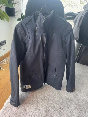 Mörkblå jacka från G-Star RAW - Snygg mörkblå jacka från G-Star RAW med huva, dragkedja och flera fickor. Jackan har en sportig look med detaljer som tryckknappar och logopatch nertill. Perfekt för dig som vill ha en stilren och funktionell jacka till vardags.