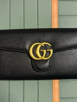 Svart kuvertväska från Gucci - Snygg svart kuvertväska från Gucci i äkta läder med guldfärgad GG-logga framtill. Väskan har en elegant kedjerem i guld och en stilren, avlång form. Perfekt accessoar för dig som gillar lyxiga detaljer och klassisk design.