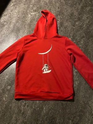 Röd hoodie med en astronaut som gungar på månen. - Cool röd hoodie med huva och ett unikt tryck av en astronaut som gungar från en måne på bröstet. Hoodien har lång ärm och är tillverkad i mjukt material, perfekt för en avslappnad och trendig stil.