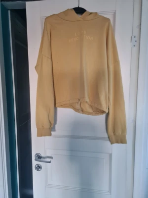 Gul cropped hoodie, Love Revolution - Säljer en gul cropped hoodie från Love Revolution med huva och långa ärmar. Tröjan har tryckt text framtill och breda muddar vid ärmsluten. Perfekt för en avslappnad och trendig look.