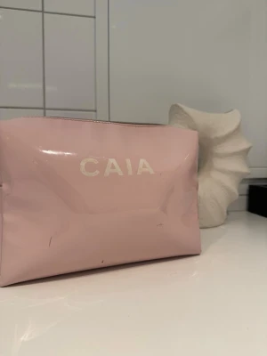 Rosa necessär från CAIA - Söt rosa necessär från CAIA i glansigt material med dragkedja upptill. Perfekt för smink eller småprylar. Enkel och stilren design med CAIA-logga i vitt på framsidan. Smidig att ha med i väskan eller på resan.