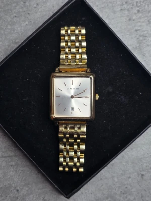 Guldfärgad fyrkantig klocka Carlheim - Stilren klocka från Carlheim med fyrkantig urtavla i silver och guldfärgat metallarmband. Urtavlan har minimalistiska markeringar och datumvisning. Perfekt accessoar för dig som gillar klassisk och elegant design.