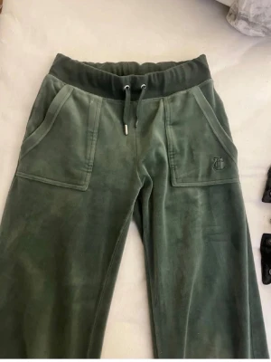 Gröna velour mjukisbyxor med fickor - Mörkgröna mjukisbyxor i mjuk velour med bred resårmidja och dragsko. Stora fickor fram och bakfickor med lock och silverknappar. Broderad logga på ena fickan fram. Perfekta för chill dagar och riktigt sköna att ha på sig.