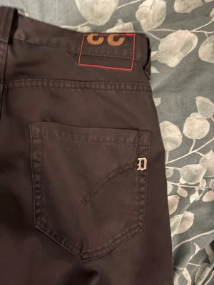 Svarta dondup jeans storlek 32 34 - Säljer ett par svarta jeans från Dolce & Gabbana med raka ben och klassisk femficksdesign. Storlek 32 34 Jeansen har en metallogo på bakfickan och en snygg detalj med röd söm vid linningen. Perfekta för dig som gillar stilrena och exklusiva plagg.