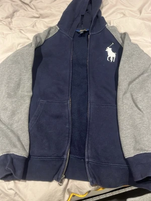 Blå och grå hoodie Polo Ralph Lauren - Mörkblå hoodie med grå ärmar från Polo Ralph Lauren. Tröjan har dragkedja, huva och stora fickor framtill. På bröstet finns det klassiska vita Polo-logotrycket med häst och ryttare. Perfekt för en avslappnad och sportig stil. Vid snabbaffär kan priset sänkas. Storlek 13-15 år