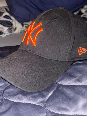 Svart New Era keps med orange NY-logga - Säljer en svart keps från New Era med böjd skärm och broderad orange NY-logga framtill. Kepsen har även en orange New Era-logga på sidan. Klassisk basebollmodell i bomull, perfekt för en sportig och trendig look.